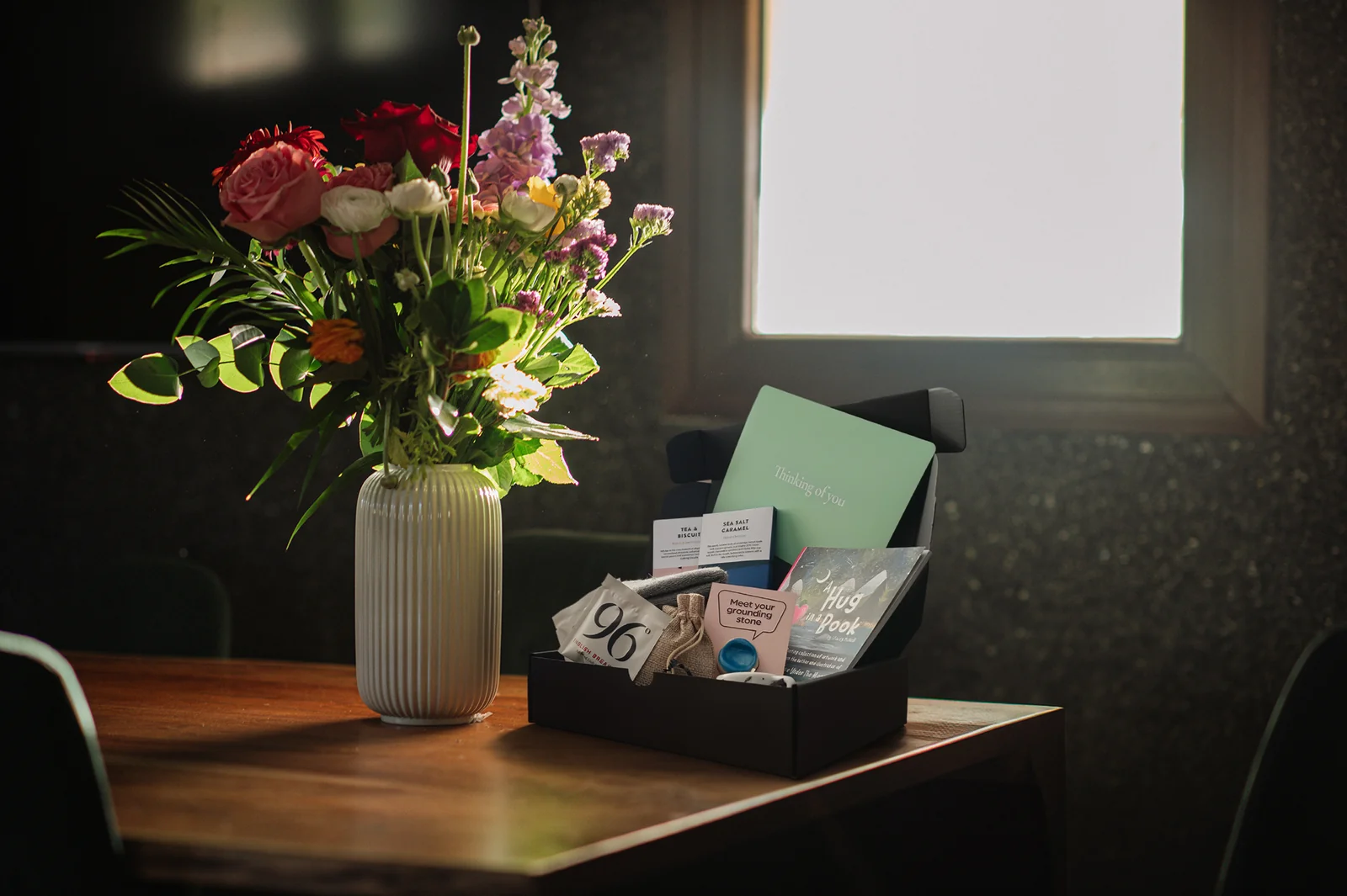 The Little Box of Mindfulness gift box open on a table beside a vase of flowers, with a 'Thinking of you' journal, tea, chocolates and the A Hug in a Book collection inside